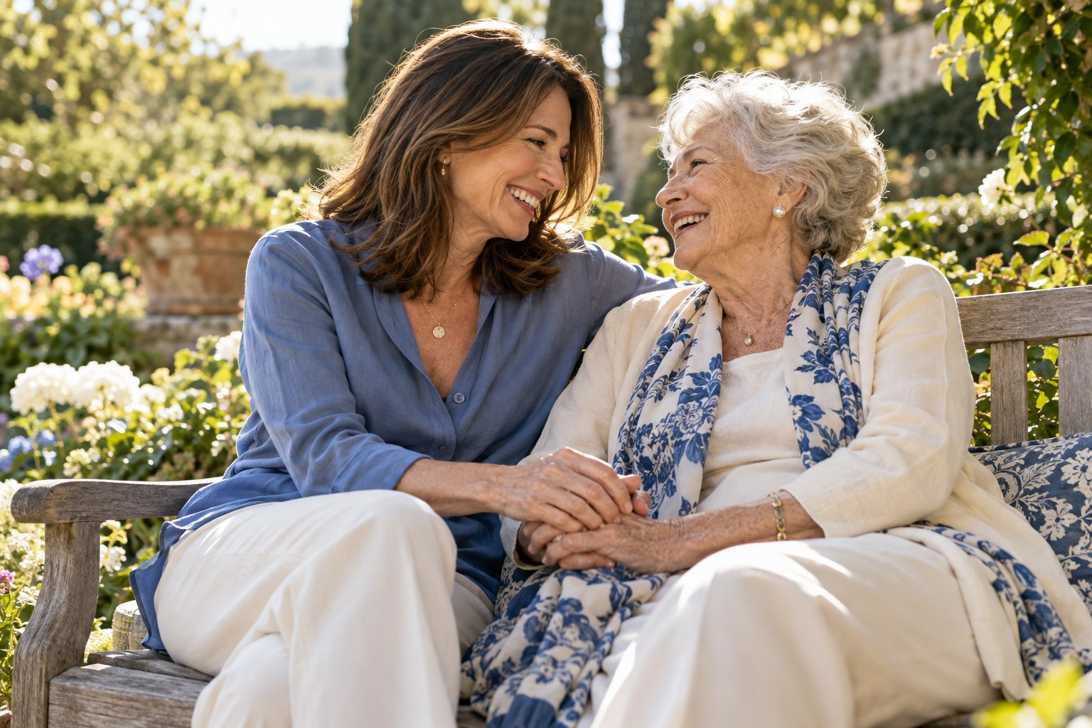An adult daughter and her elderly mother laughing together and holding hands on a garden bench